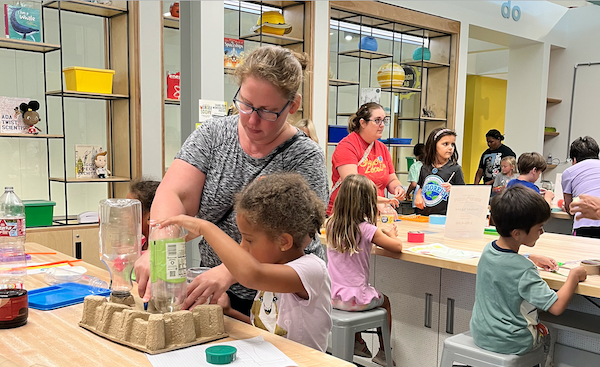 A woman and child work together at a table in a brightly lit space, using recycled packaging and plastic bottles to build.