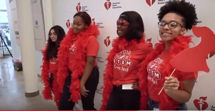 Four teen girls wearing red STEM T-shirts pose with red feather boas in front of a step and repeat banner