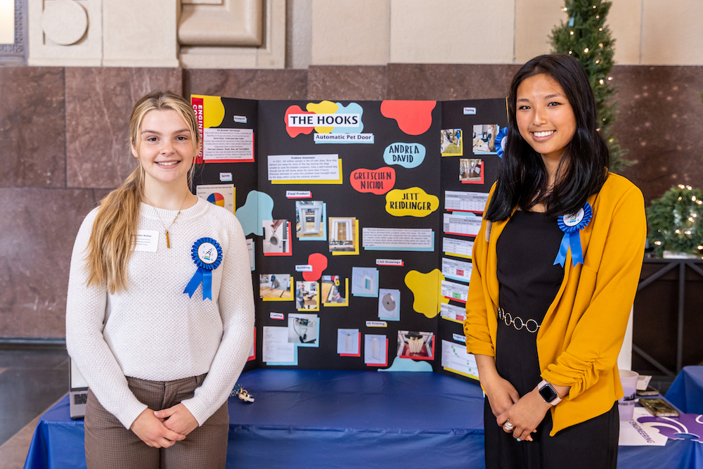 Two female students stand before a colorful project display board.