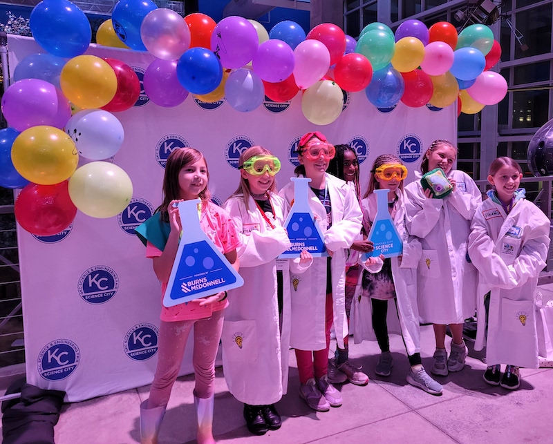 A group of five girls wearing lab coats and holding chemistry signs smile as they pose in front of a balloon-topped backdrop