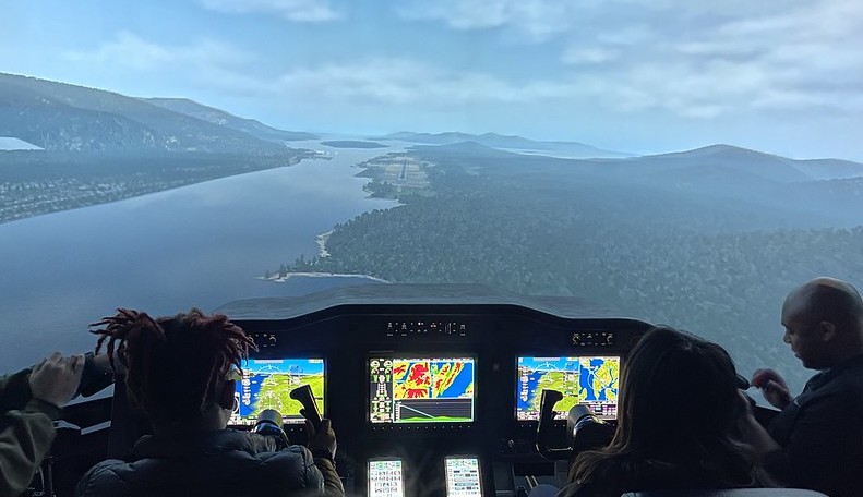 Pictured from behind are two middle school girls at the controls of an aircraft simulator, looking into a screen depicting a lake and sky the plane is flying over.