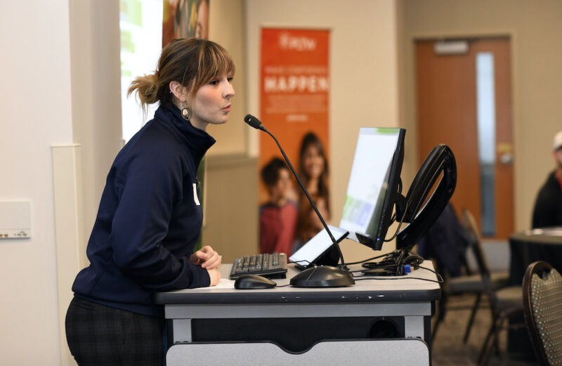 Allison Nelson is talking into a microphone on a podium, with an orange PLTW banner in the background.
