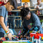 Two elementary school-age boys focus on their LEGO robot, which is placed on a colorful game mat with other LEGO components.