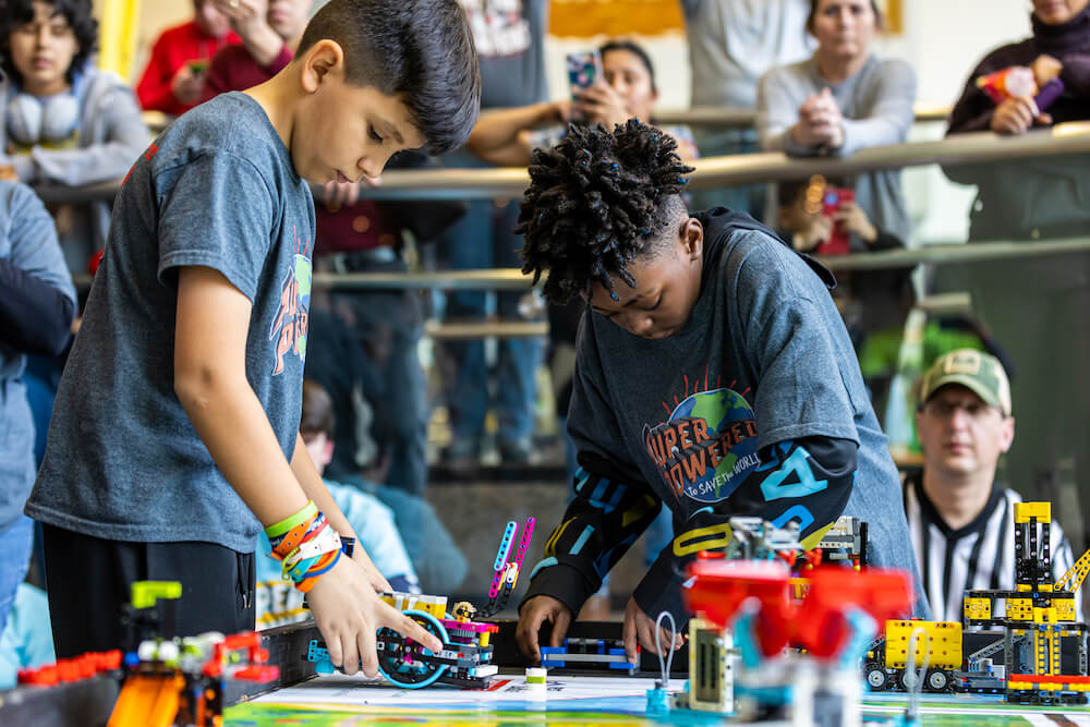 Two elementary school-age boys focus on their LEGO robot, which is placed on a colorful game mat with other LEGO components.